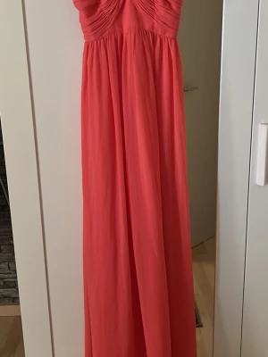 Lång korallrosa klänning med drapering - Snygg långklänning i korallrosa med draperad överdel och smala axelband. Klänningen har en flowy passform och markerad byst, perfekt för sommarens tillfällen. Materialet känns lätt och luftigt.
