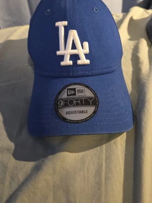 Blå LA-keps från New Era 9FORTY - Snygg blå keps från New Era, modell 9FORTY, med justerbar passform. Framtill sitter ett broderat LA-logotyp i vitt och på sidan finns New Era-loggan. Klassisk böjd skärm och ventilerande hål upptill. Perfekt för dig som gillar streetwear och sportig stil.