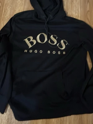Svart hoodie från Hugo Boss - Snygg svart hoodie från Hugo Boss med stor logga i vitt och guld på bröstet. Klassisk huva med dragsko och känguruficka framtill. Perfekt för en stilren och avslappnad look. Materialet känns mjukt och bekvämt. Den är limted edition så först till kvarn!