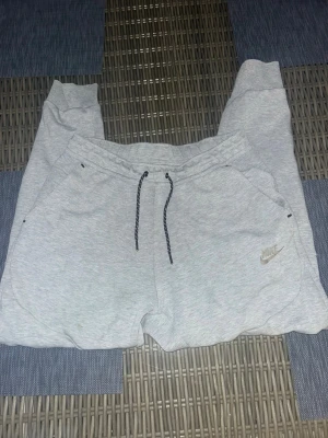 Gråa mjukisbyxor från Nike - Ljusgrå mjukisbyxor från Nike med resår i midjan och svarta dragsnören. Byxorna har två sidofickor och en bakficka med dragkedja. Nike-logga broderad på vänster ben. Perfekta för chill eller träning.Ända defekten är att märket tappat färg 