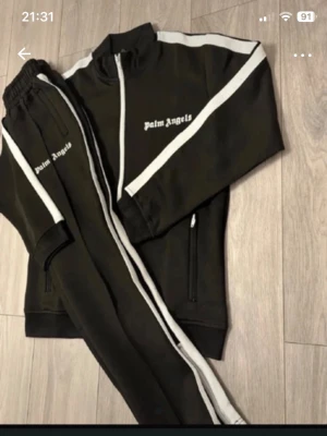 Palm Angels svarta mjukisbyxor - Palm Angels svarta mjukisbyxor med vita sidoränder och logga tryckt på benet. Klassisk sportig look med resår i midjan och raka ben. Perfekta för en avslappnad och trendig stil.