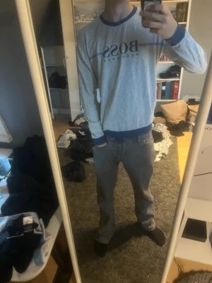 Grå långärmad tröja från Hugo Boss - Snygg grå långärmad tröja från Hugo Boss med blå detaljer vid hals, ärmslut och midja. Tröjan har tryckt logga och tunna blå linjer över bröstet. Perfekt för en avslappnad och stilren look.