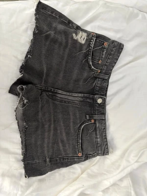 Svarta jeansshorts med rå kant - Säljer ett par svarta jeansshorts med råa, fransiga kanter och slitningar framtill. Shortsen har klassisk femficksdesign, knappgylf och bälteshällor. Perfekta för en avslappnad stil.