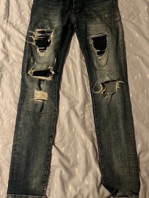 Blå slitna jeans med stora hål - Säljer ett par blå jeans med kraftiga slitningar och stora hål på båda benen. Jeansen har en rak passform och klassisk femficksdesign. Perfekta för dig som gillar en edgy och trendig streetstil.