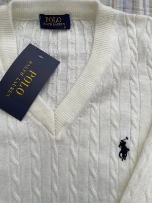 Vit kabelstickad tröja Polo Ralph Lauren - Kabelstickad vit tröja från Polo Ralph Lauren med V-ringning och klassisk broderad logga i svart på bröstet. Långa ärmar och ribbade muddar ger en clean och tidlös look. Perfekt till jeans eller kjol för en preppy stil.