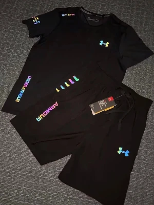 Svart träningsset från Under Armour - Säljer ett par svart träningsset från Under Armour med färgglada loggor och detaljer. Shortsen har snörning i midjan och är tillverkade i ett lätt och sportigt material som passar perfekt för träning. Stilen är modern och loggorna syns tydligt på ena benet. Storlek S, helt ny