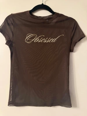 Brun mesh t-shirt med text - Snygg brun mesh t-shirt med korta ärmar och texten 'Obsessed' i vit skrivstil på bröstet. T-shirten är genomskinlig och har en tight passform