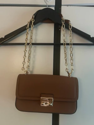 Michael Kors Axelväska - Snygg brun handväska från Michael Kors i slätt läder. Väskan har en rektangulär form, guldfärgad kedjerem och ett elegant metallspänne framtill med logga. Perfekt accessoar för att lyfta din outfit. Michael Kors axelväska i cognacsfärg. Aldrig använd. Köpt pă NK. Original dustbag medföljer. Väskan är i nyskick och saljs eftersom den inte kommit till användning. Köpt pă NK. Mått: Botten: 6,5 cm Höjd: 14 cm Bredd: 22 cm Axelbandet: ca 124 Guld kedja 47+47 Läder del 15+15