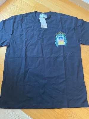 Svart t-shirt från Casablanca - Snygg svart t-shirt från Casablanca med färgglad tryckt grafik både fram och bak. På bröstet står det 'Avenida Casablanca' och på ryggen finns ett stort motiv av ett fönster med utsikt mot en byggnad och gröna blad. Klassisk passform och rund hals. Perfekt för dig som gillar streetwear.