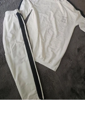 Vitt loungewear-set med strassdetaljer - Snyggt vitt loungewear-set med svarta ränder längs ärmar och byxben. Tröjan har små strassdetaljer över hela framsidan och ett stort H i strass på bröstet. Mjukt och bekvämt material, perfekt för chill dagar hemma.