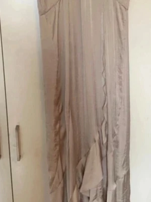 Champagnefärgad långklänning med volang - Elegant långklänning i champagnefärgad satin med vackra volangdetaljer framtill. Klänningen har en mjuk, glansig yta och ett svepande fall som ger en lyxig känsla. Perfekt för dig som vill sticka ut med en stilren och trendig look.