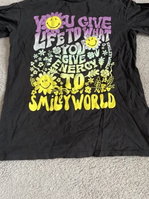 Svart SmileyWorld t-shirt med tryck - Cool svart t-shirt från SmileyWorld i storlek 152 med färgglatt tryck i lila, vitt och gult framtill. Texten och blommorna ger en retrovibe och flera smileys pryder framsidan. Klassisk passform och korta ärmar, perfekt för dig som gillar statement-plagg.