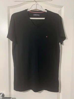 Svart v-ringad t-shirt Tommy Hilfiger - Klassisk svart t-shirt från Tommy Hilfiger med v-ringning och liten broderad logga på bröstet. Tillverkad i mjuk bomull och har korta ärmar. Perfekt basplagg med stilren design och diskreta detaljer.Finns att hämta i Södertälje.