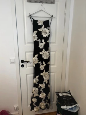 Svart långklänning med blommor - Elegant svart långklänning med stora vita blommor och smala axelband. Klänningen har en figurnära passform och är perfekt för speciella tillfällen. Materialet känns mjukt och följsamt mot kroppen.