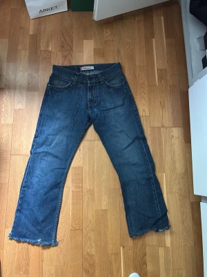 Levi's blå raka jeans med rå kant - Säljer ett par klassiska blå Levi's jeans med rak passform och rå, fransad kant nedtill. Jeansen har fem fickor och normal midja. Tillverkade i slitstark denim som ger en tidlös look. Perfekta för dig som gillar en avslappnad och trendig stil.