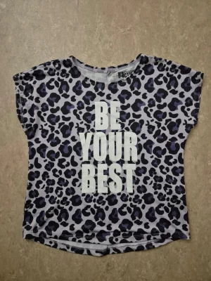 T-shirt - Cool t-shirt från H&M med leopardmönster i svart och lila på ljus bakgrund. Stor vit text 'BE YOUR BEST' framtill. Kortärmad modell i mjukt material, perfekt för en avslappnad och trendig look.