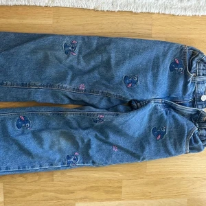 Blå jeans med Stitch-broderi - Säljer ett par blå jeans med broderade Stitch-motiv och små rosa tassavtryck. Jeansen har resår i midjan bak och raka ben. Perfekta för dig som gillar Disney och vill ha något unikt i garderoben!