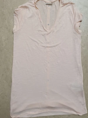 Blus - Ljusrosa t-shirt i tunn bomull med korta ärmar och v-ringning. Knappar längs hela ryggen ger en unik detalj. Lätt transparent och loose fit för en avslappnad look. Perfekt till jeans eller kjol.