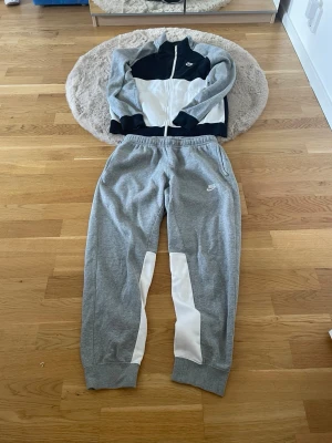 Nike grå och svart träningsset - Nike träningsset med zip-jacka och byxor. Jackan är svart, vit och grå med dragkedja och hög krage, medan byxorna är grå med vita detaljer och resår i midjan. Båda delarna har Nike-logga och är tillverkade i mjukt material, perfekt för träning eller chill.