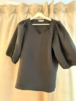 Svart kort klänning med puffärm - Stilren svart kort klänning från Indiska med v-ringning och voluminösa puffärmar. Klänningen har en enkel, rak passform och är tillverkad i ett strukturerat tyg som ger en snygg känsla. Perfekt för dig som gillar minimalistisk stil.