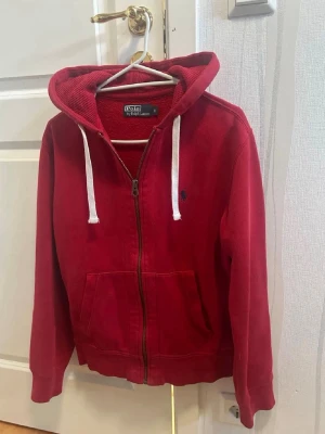 Röd hoodie från Ralph Lauren - Snygg röd hoodie från Ralph Lauren med dragkedja, kängurufickor och vit dragsko i huvan. Klassisk blå logga broderad på bröstet. Mjuk bomullskvalitet och ribbade muddar vid ärmslut och nederkant. Perfekt för en avslappnad och stilren look.