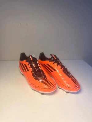 Adidas F50 orange FG (äldre modell) - Snygga Adidas F50 fotbollsskor i en skarp orange färg med svarta detaljer och klassiska tre ränder på sidan. Skorna har svarta snören och vit hälkappa. Tillverkade i syntetmaterial för lätt vikt och bra bollkontroll. Perfekta för dig som vill sticka ut på planen.