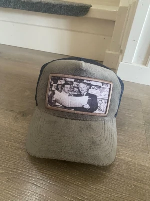 Grå och marinblå truckerkeps - Säljer en grå och marinblå truckerkeps med mesh på sidorna och baksidan. Framtill finns en stor patch med svartvitt fototryck av personer och dollarbakgrund. Kepsen har böjd skärm och justerbar snapback. Perfekt för en avslappnad streetwear-look. Den svarta kepsen kostade 499kr ny och den gråa typ 900. Båda kepsarna för endast 399kr