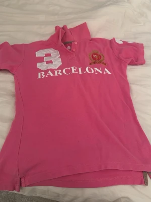 Rosa pikétröja Barcelona Designs - Säljer en rosa pikétröja från Barcelona Designs med tryckta detaljer som siffran 3 och texten 'Barcelona' på framsidan. Tröjan har korta ärmar, krage och broderat emblem på bröstet. Perfekt för dig som gillar sportig stil.