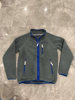 Grå fleecejacka från Patagonia - Säljer en grå fleecejacka från Patagonia med blå detaljer och dragkedjor. Jackan har tre fickor med dragkedja och hög krage. Perfekt för lager-på-lager och outdoor. Materialet är mjuk fleece som håller dig varm under kyliga dagar.