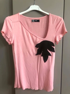 Rosa t-shirt från ONLY med svart detalj - Snygg rosa t-shirt från ONLY med korta ärmar och en unik svart bladapplikation på bröstet. T-shirten har en djup v-ringning och vågiga kanter vid ärmslut och nederkant. Perfekt för dig som vill ha något enkelt men ändå speciellt.