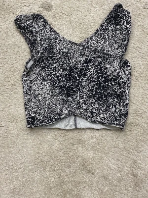Svartvit croppad topp med mönster - Säljer en croppad topp i svart och vitt med abstrakt mönster. Toppen har breda axelband och en omlottliknande nederkant. Perfekt för dig som gillar en edgy stil och vill sticka ut lite extra.