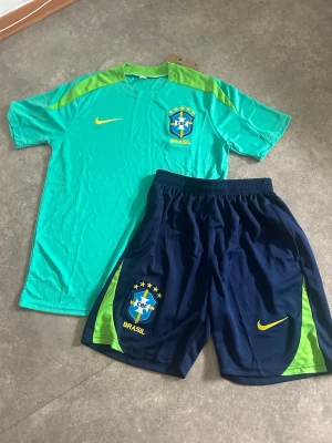Brasilien Nike fotbollsträningsset - Snyggt träningsset från Nike med Brasilien-tema. T-shirten är turkos med limegröna detaljer och Brasilien-logga på bröstet. Shortsen är mörkblå med limegröna inslag och har även Brasilien-logga. Perfekt för fotbollsträning eller sport. Helt oanvänd 