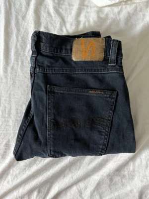 Svarta/väldigt mörkblåa lite tajtare jeans från Nudie Jeans - Säljer ett par svarta/väldigt mörka jeans från Nudie Jeans med klassisk femficksmodell och läderpatch med orange logga bak. Jeansen har smal passform och är tillverkade i mjuk denim med stretch för extra komfort.