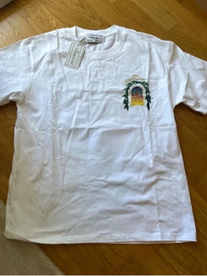 Vit Casablanca t-shirt med tryck - Säljer en vit t-shirt från Casablanca med färgglatt tryck av en byggnad och texten 'Avenida Casablanca' på ryggen. T-shirten har korta ärmar och rund hals. Perfekt för dig som gillar unika prints och streetwear-vibe.