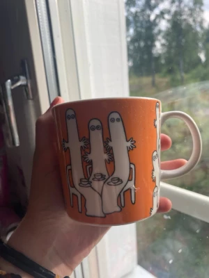 Muminmugg med orange motiv - En orange mugg med motiv av Hattifnattar från Mumin. Muggen har ett vitt handtag och är dekorerad med flera vita figurer runt hela koppen. Perfekt för te eller kaffe och en fin samlardetalj för Mumin-fans.