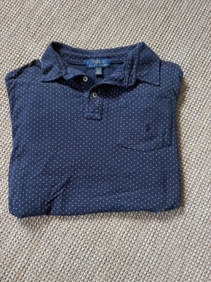 Mörkblå prickig Polo Ralph Lauren - Snygg mörkblå  pique från Polo Ralph Lauren med vita prickar, klassisk krage och bröstficka med broderad logga. Piquen har knappar framtill och är tillverkad i mjuk bomull. Perfekt till golfbanan eller bara vara snygg nu till sommaren 
