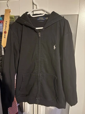 Svart hoodie från Polo Ralph Lauren - Svart hoodie med dragkedja från Polo Ralph Lauren i modellen Custom Fit. Klassisk design med huva, två stora fickor framtill och den ikoniska vita logon broderad på bröstet. Perfekt för en avslappnad och stilren look.