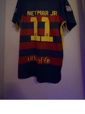 Barcelona Neymar JR matchtröja - Säljer en FC Barcelona matchtröja med Neymar JR och nummer 11 på ryggen. Tröjan är blå med röda horisontella ränder, gula detaljer och klubbmärke på bröstet. Tillverkad i lätt och ventilerande material, perfekt för fotboll eller som supporterplagg. Storlek S