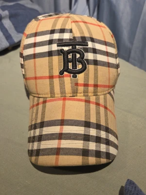 Beige rutig keps från Burberry - Säljer en beige keps från Burberry med klassiskt rutigt mönster i svart, vitt och rött. Kepsen har en böjd skärm och ett broderat svart TB-monogram framtill. Justerbar rem baktill för perfekt passform. Tillverkad i bomull.