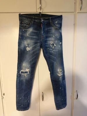 Blå Dsquared2 jeans med slitningar - Säljer ett par blå Dsquared2 jeans i storlek 44 med coola slitningar och färgstänk. Jeansen har klassisk femficksmodell, raka ben och snyggt läderpatch bak i midjan. Perfekta för dig som gillar streetstyle och trendiga detaljer.