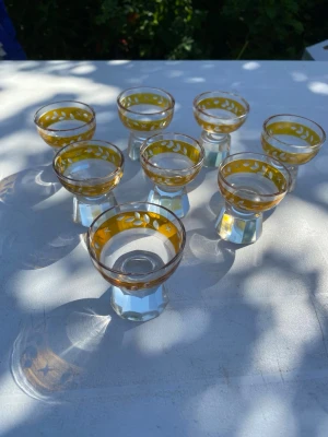 Set med 7 små glas med guldkant - Säljer ett set med sju små glas med guldfärgad dekor och mönster av blad och stjärnor. Glasen har en sexkantig fot och är tillverkade i klart glas med detaljer i gult och vitt. Perfekta för snaps eller likör. Troligtvis Hofmantorp skiljer sig lite i storlek, 3 stycken har nagg. 