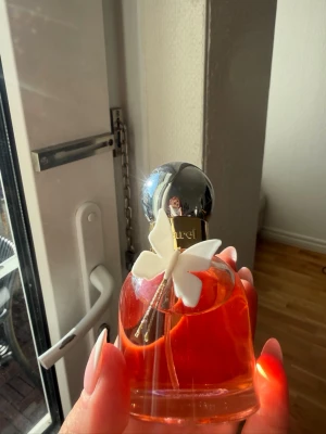 Cacharel Ella Ella 50ml - Elegant parfym från Cacharel i en rund glasflaska med orange doftvätska. Flaskan har en silverfärgad rund kork och dekoreras med en vit blomma på framsidan. Rymmer 50 ml och har en lyxig och feminin design som sticker ut i badrumshyllan. Använd ungefärg en gång