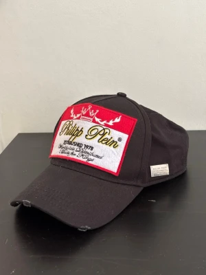 Svart keps från Philipp Plein - Snygg svart keps från Philipp Plein med stor broderad patch framtill i rött, vitt och gul.  Kepsen har böjd skärm och justerbar rem bak. Märkesdetalj på sidan.