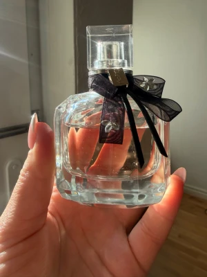 Ysl Mon Paris 50ml - Säljer en elegant parfym från Lancôme, La Vie Est Belle. Flaskan är i tjockt, genomskinligt glas med en fyrkantig form och har en söt svart rosett med dekorativa detaljer runt halsen. Locket är transparent och flaskan känns lyxig och tidlös. 