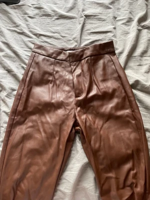 Bruna byxor i fuskskinn med hög midja - Snygga bruna byxor i fuskskinn med hög midja och raka ben. Byxorna har pressveck framtill och en ficka bak. Perfekta för dig som vill ha en trendig och stilren look.