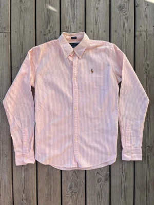 Rosa och vit randig skjorta Polo Ralph Lauren - Den perfekta skjortan till sommaren i en rosa randig modell. Storlek står 6 men motsvarar en slim fit S.Klassisk långärmad skjorta från Polo Ralph Lauren i rosa och vitt randigt bomullstyg. Skjortan har knappar framtill, vet ej om det är herr eller dam, button-down krage och den ikoniska broderade loggan på bröstet. Perfekt för en stilren och avslappnad look