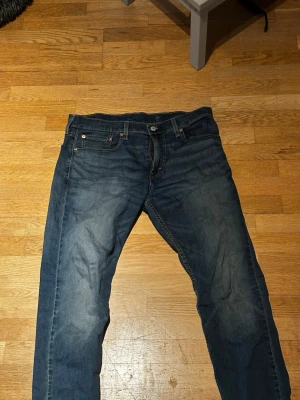 Levi's blå raka jeans - Säljer ett par klassiska blå jeans från Levi's med rak passform och fem fickor. Jeansen har snygga slitningar framtill och tydliga sömmar på bakfickorna. Tillverkade i slitstark denim med knappgylf och läderpatch bak.