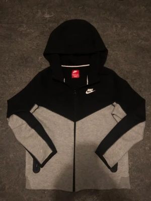 Svart Och Grå Nike Tech - Snygg Nike tech i svart och grått med dragkedja framtill och vit Nike-logga på bröstet. Storleken är XL i mindre storlek så den passar också S! Perfekt för en sportig och avslappnad stil.