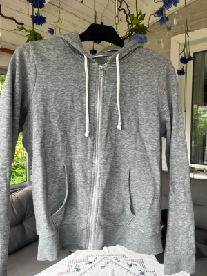 Grå zip hoodie  - Snygg gråmelerad hoodie med dragkedja framtill och vita dragsnören i huvan. Två fickor på magen och ribbade muddar vid ärmslut och nederkant. Perfekt för chill dagar eller när du vill ha en enkel och clean look.