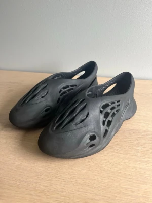 Svarta foam runners tofflor - Säljer ett par svarta foam runners tofflor. Skorna har en chunky sula och är perfekta för dig som gillar streetwear och vill sticka ut. Lätta att ta på och av, passar både hemma och ute. OBS! Har ett litet hål vid vänster stortå i skon. 